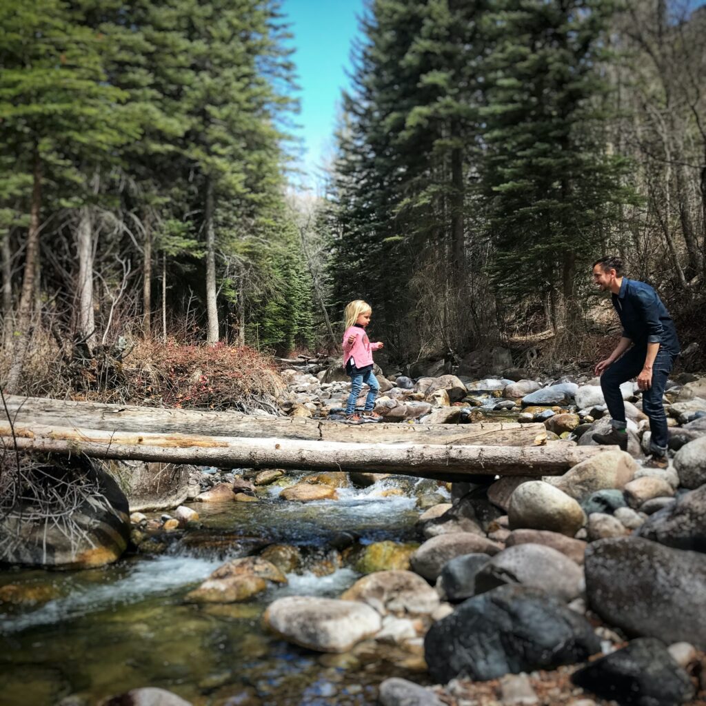 In Colorado, a little girl walks across a log over a river looking towards her father