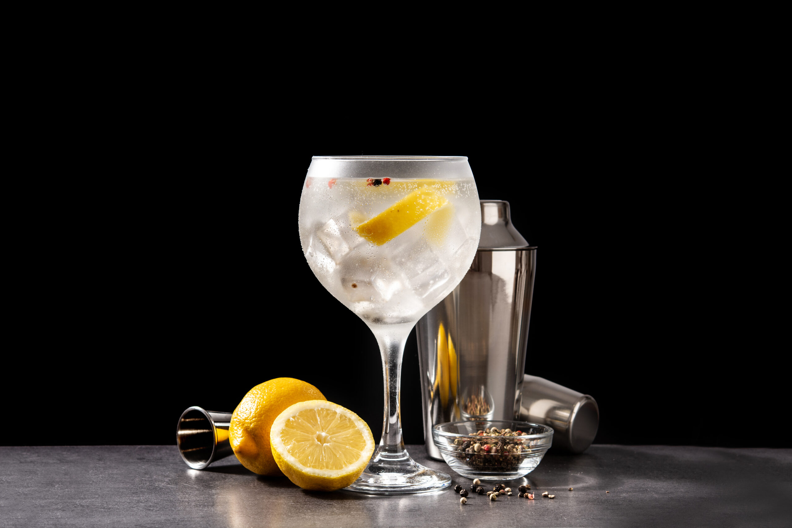 A glass filled with ice and lemon slices, with a cocktail mixer set just behind it.