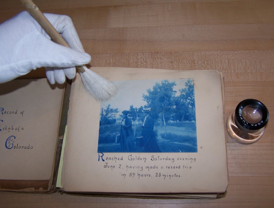 A hand with a white glove uses a brush to gently clean an old photograph