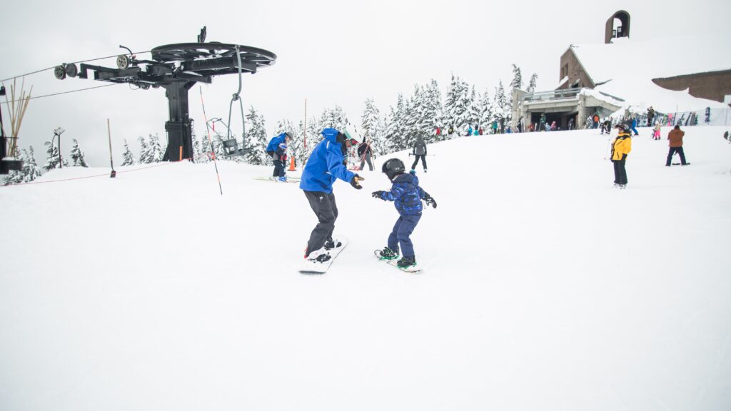 An adult assists a child who is learning to snowboard