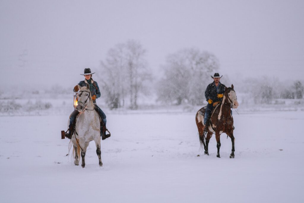 Two people wearing Stetsons, horseback riding in the snow