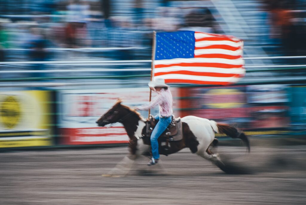 A cowboy on horseback carrying a USA flag zooms past a blurred crowd