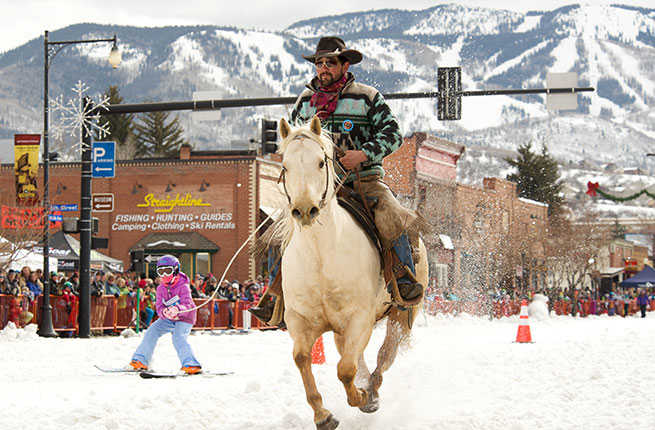 A cowboy riding a horse pulls a child on skis along a snowy road