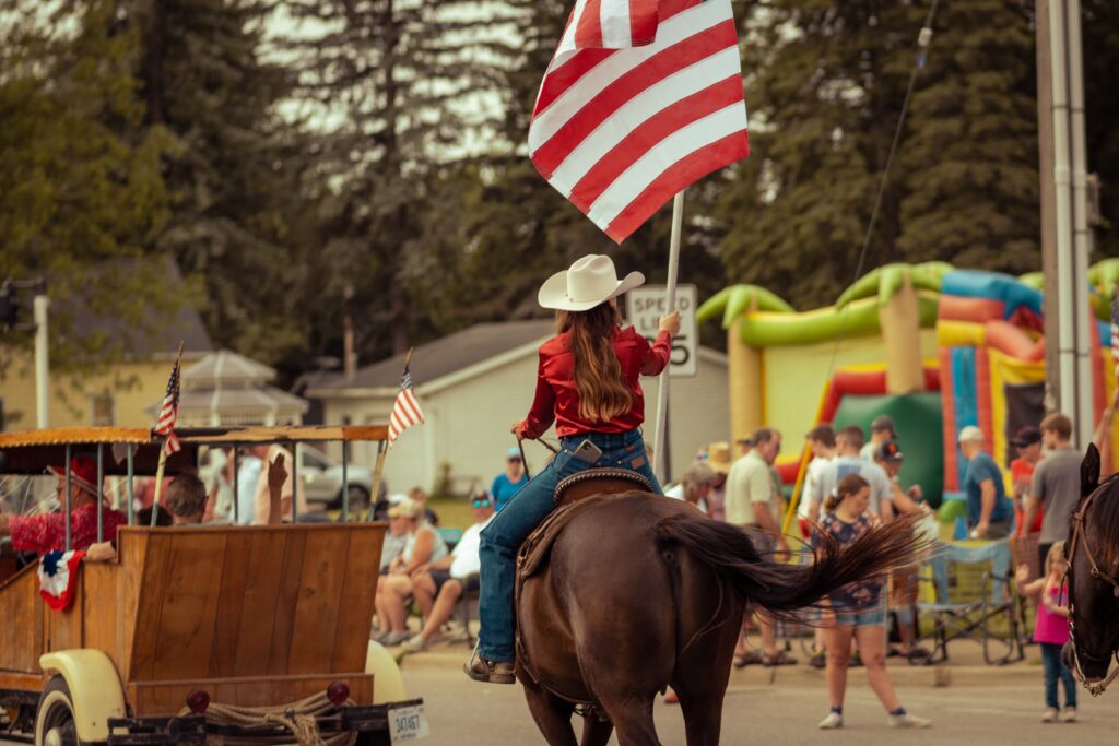 A cowgirl rides on horseback carrying the USA flag through a parade