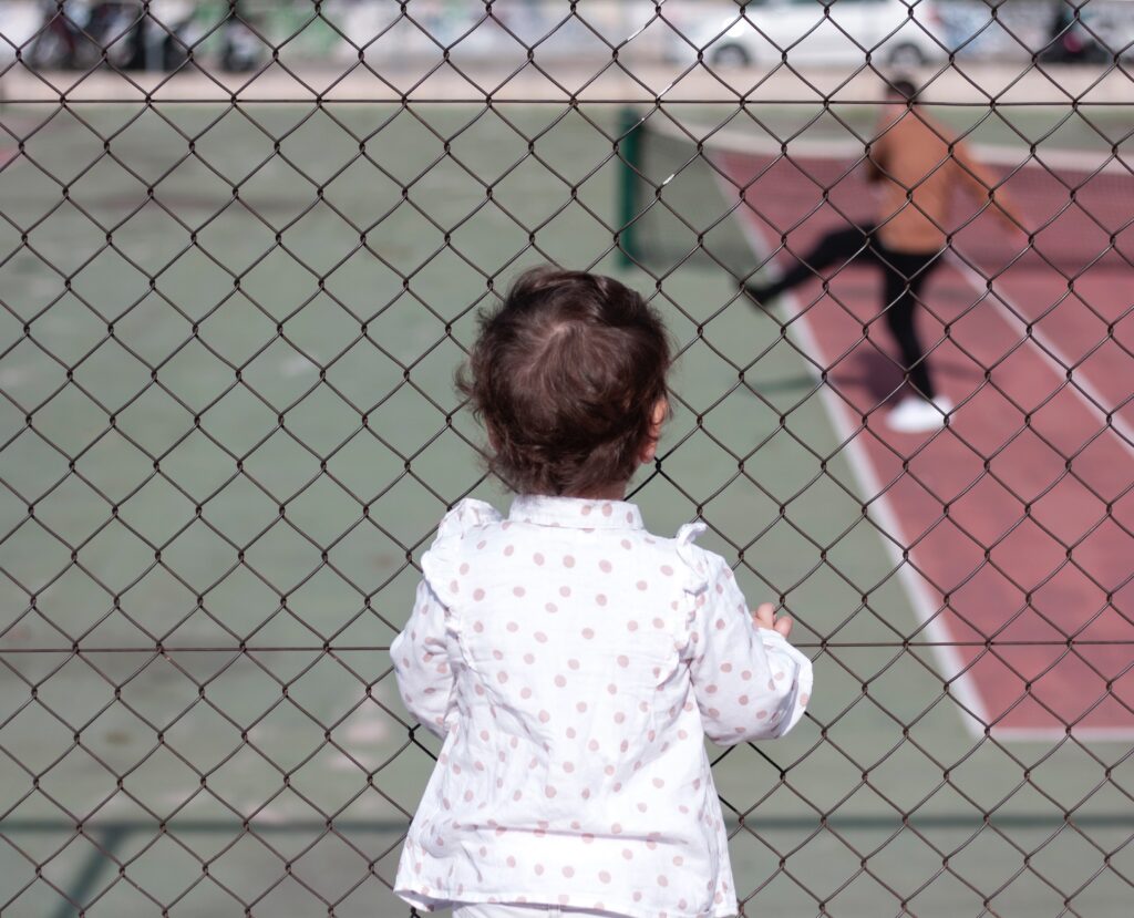 A toddler looks through a fence to a tennis court