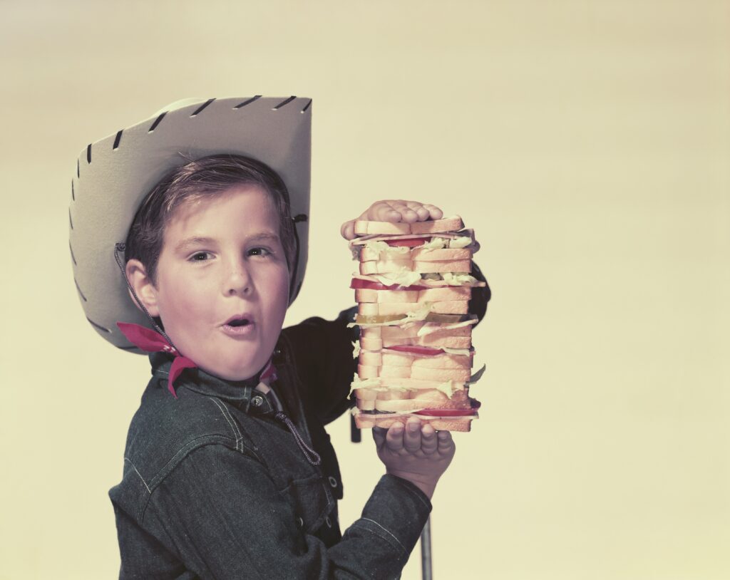 A cowboy child holding an enormous sandwich