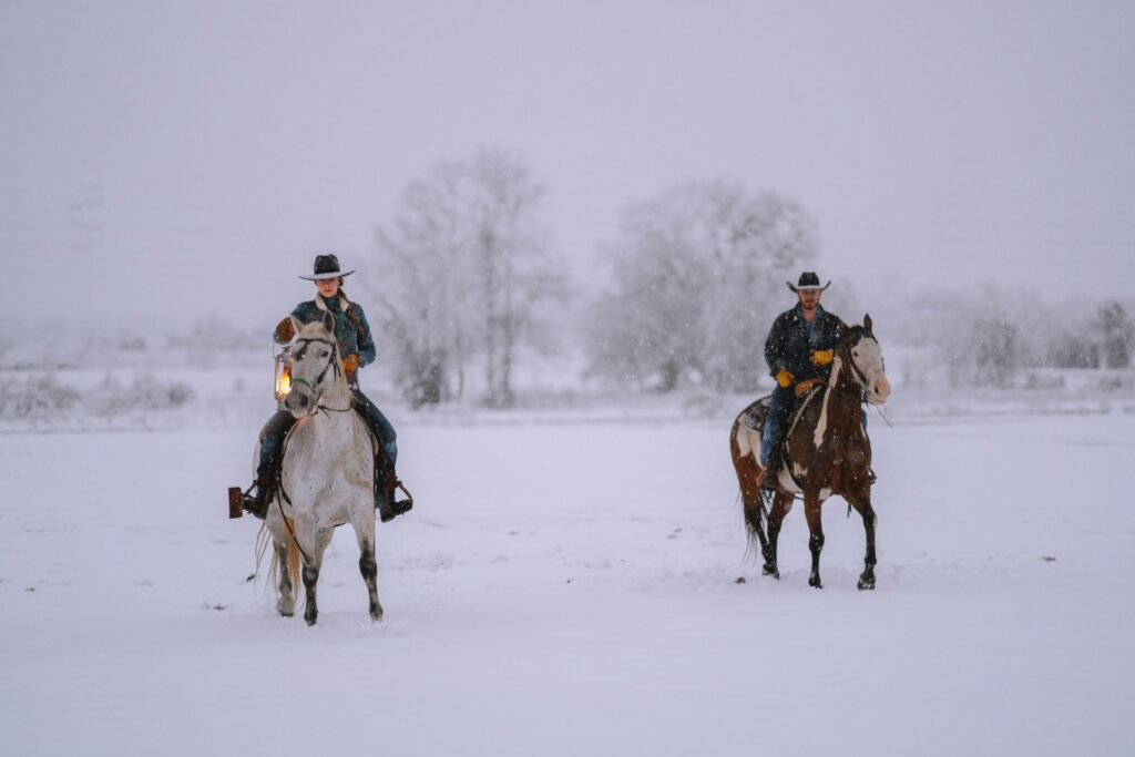 Two cowboys ride their horses through the snow