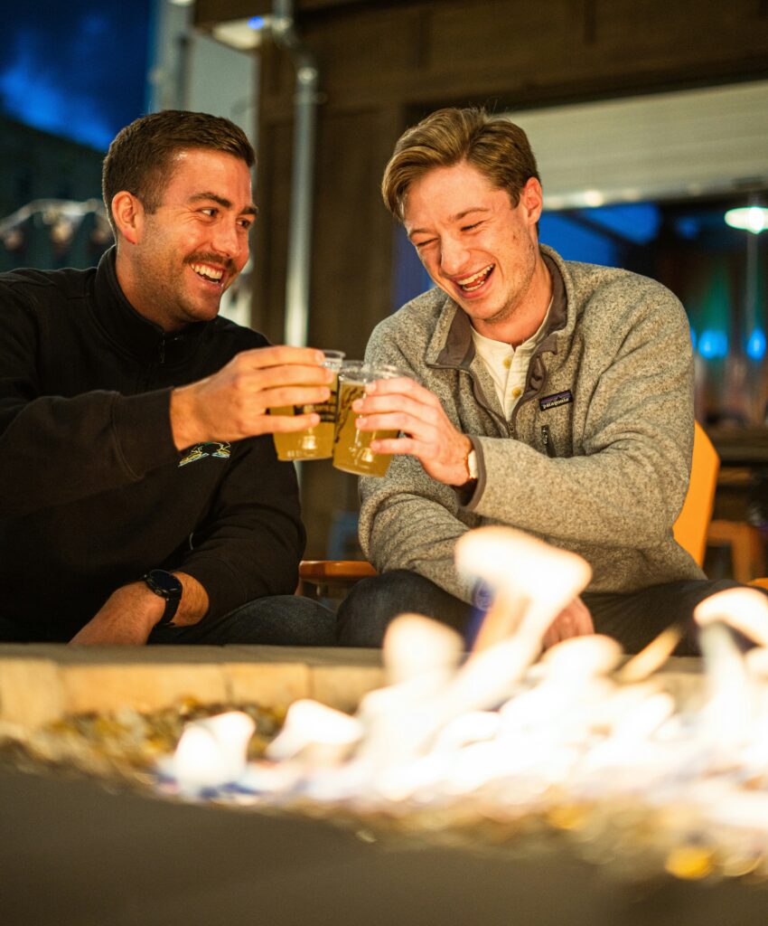 Two men laugh by a firepit whilst enjoying some beer
