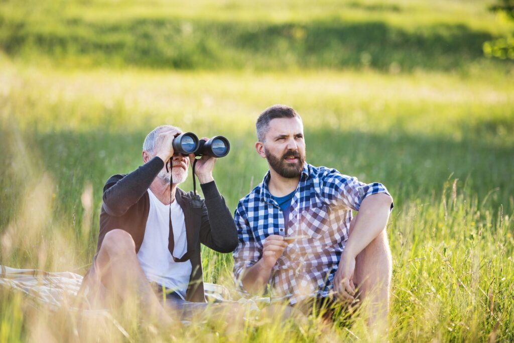 Two men sat in a field using binoculars