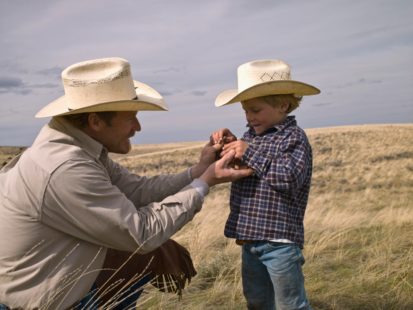 A father and a little boy holding hands and waering cowboy hats.