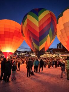 Large group of people standing around hot air balloons on the ground at night