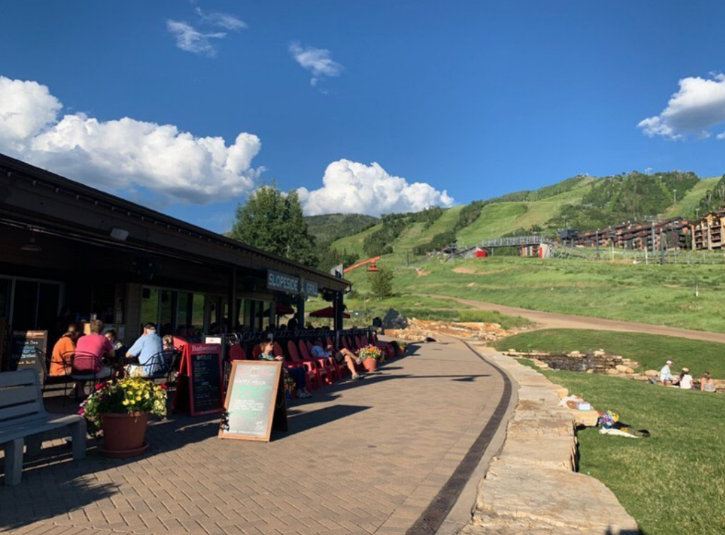 The large patio at Slopeside Grill. A view of a green Mt Werner, lots of tables with red patio chairs. 