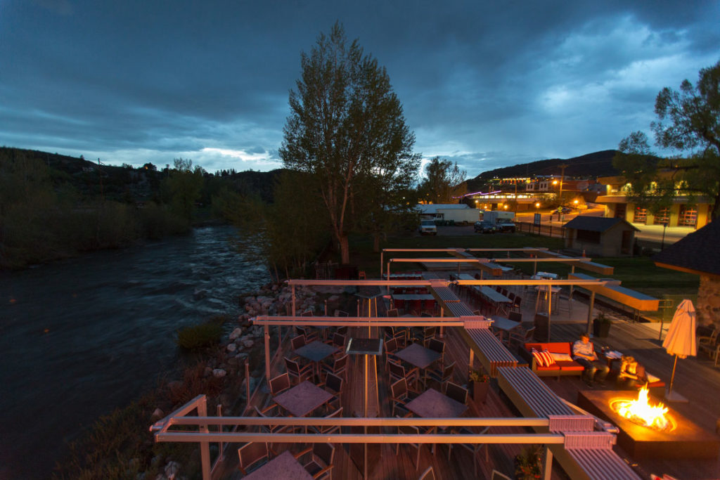 View of Aurum's patio at night, tables along the Yampa River with a fire pit and couches. 