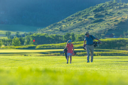 Adult man and young girl walking on a golf course carrying golf bags. Golf in Steamboat Springs is the best in Colorado.