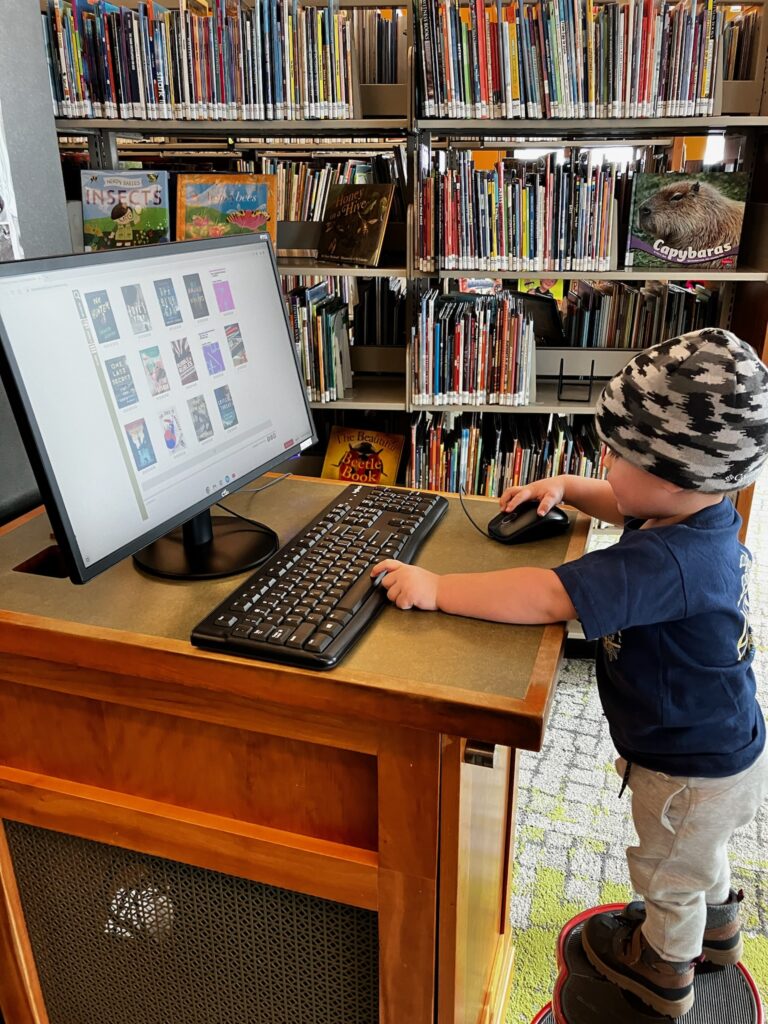 A toddler standing at a library computer and looking at the book catalogue online. 