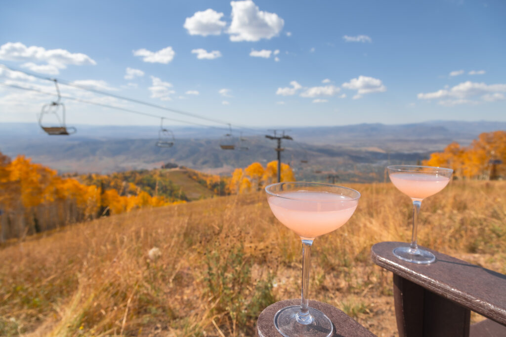 View of Mt Werner from Thunderhead Lodge. Two pick cocktails sit on wooden chair arms, overlooking the gondola and the characteristic yellow Aspen trees in Fall in Steamboat Springs.