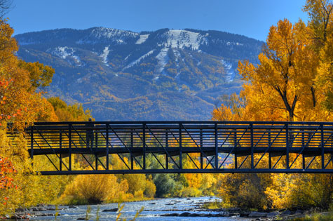 The bridge over the Yampa River with golden aspens and the rocky mountains behind part of the Core Trail hiking trail in Steamboat Springs.