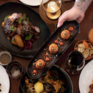 A hand holding a small plate of bite-sized appetizers over top of a table of food.