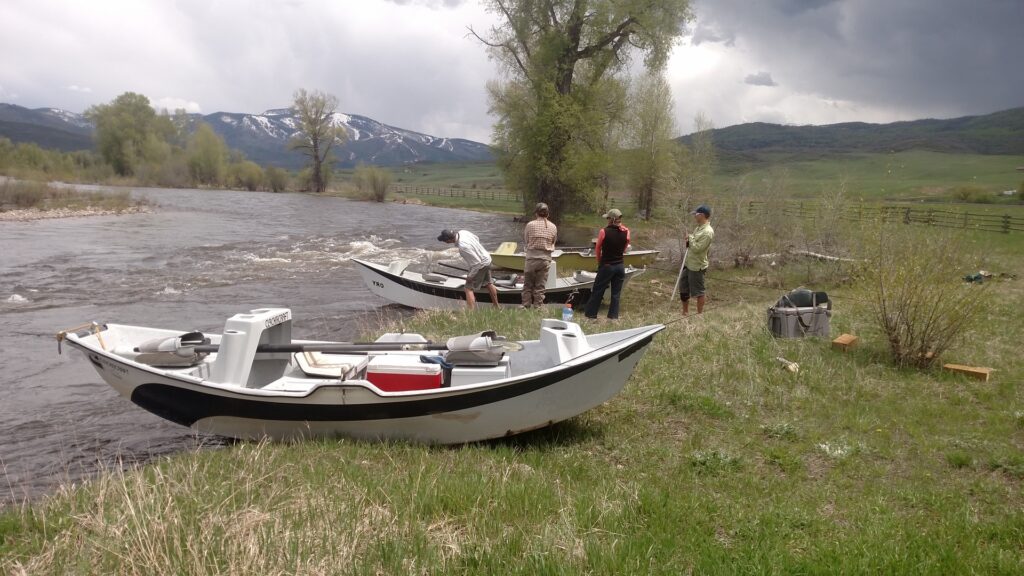 Three men fishing on the side of a river with two aluminium botas beside them.