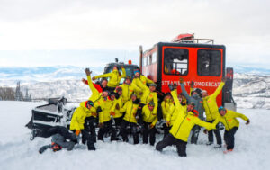 A group of skiers posing in front of the large snowcat vehicle at the top of the snowy mountain. 