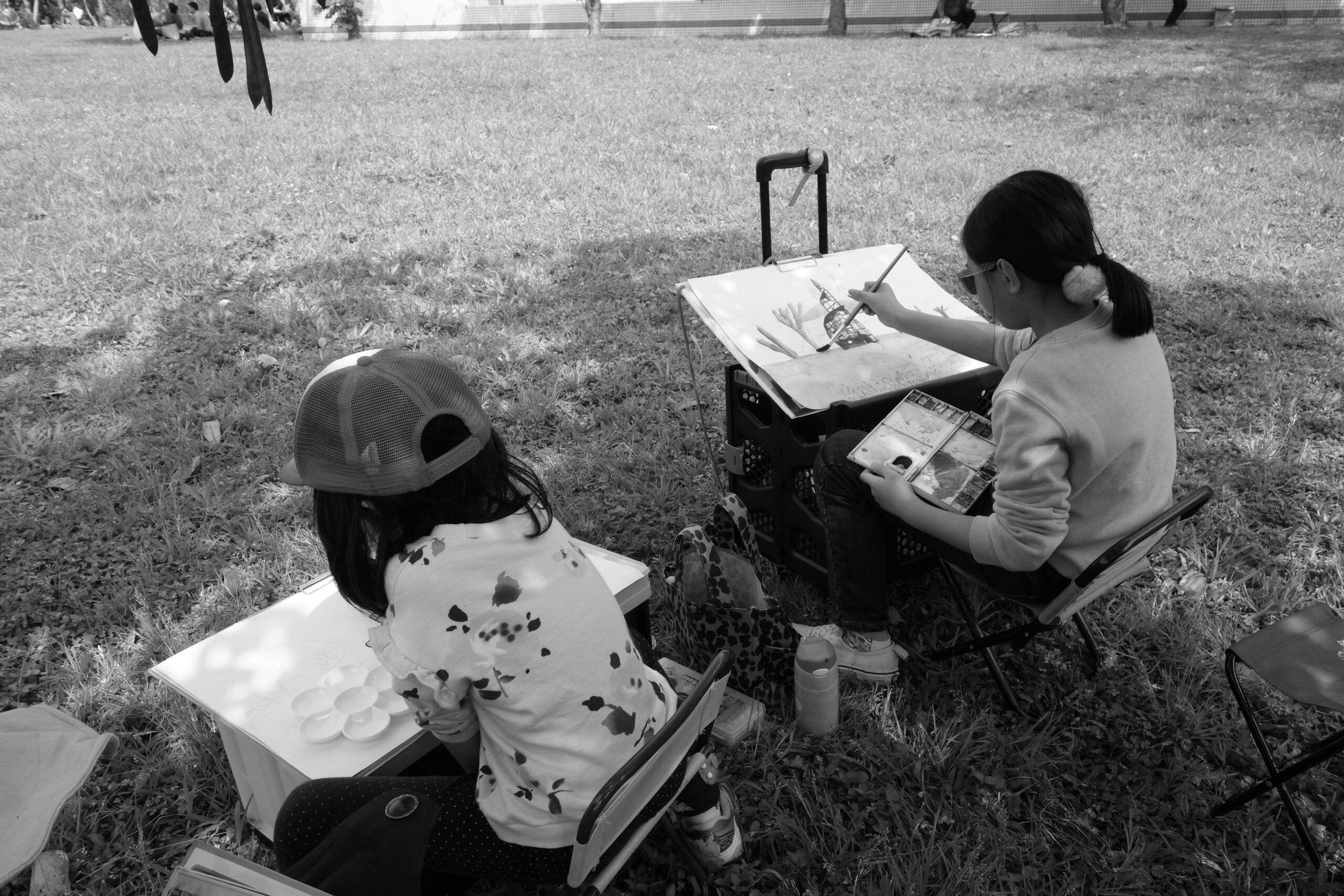 Two children sitting at easels and painting on canvas on the grass in the park.