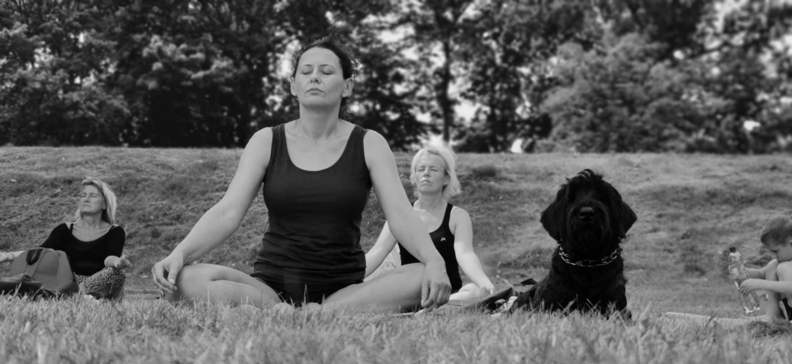 Women sitting crosslegged with black dog beside her, while doing a yoga class outdoors in a grassy area.