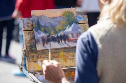 View over woman's shoulder as she paints a mountain landscape on canvas.