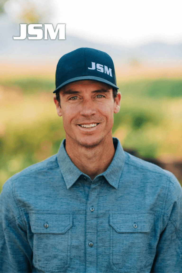 Portrait of Brian Heit, Vice President of JSM, smiling outdoors wearing a blue shirt and JSM hat.