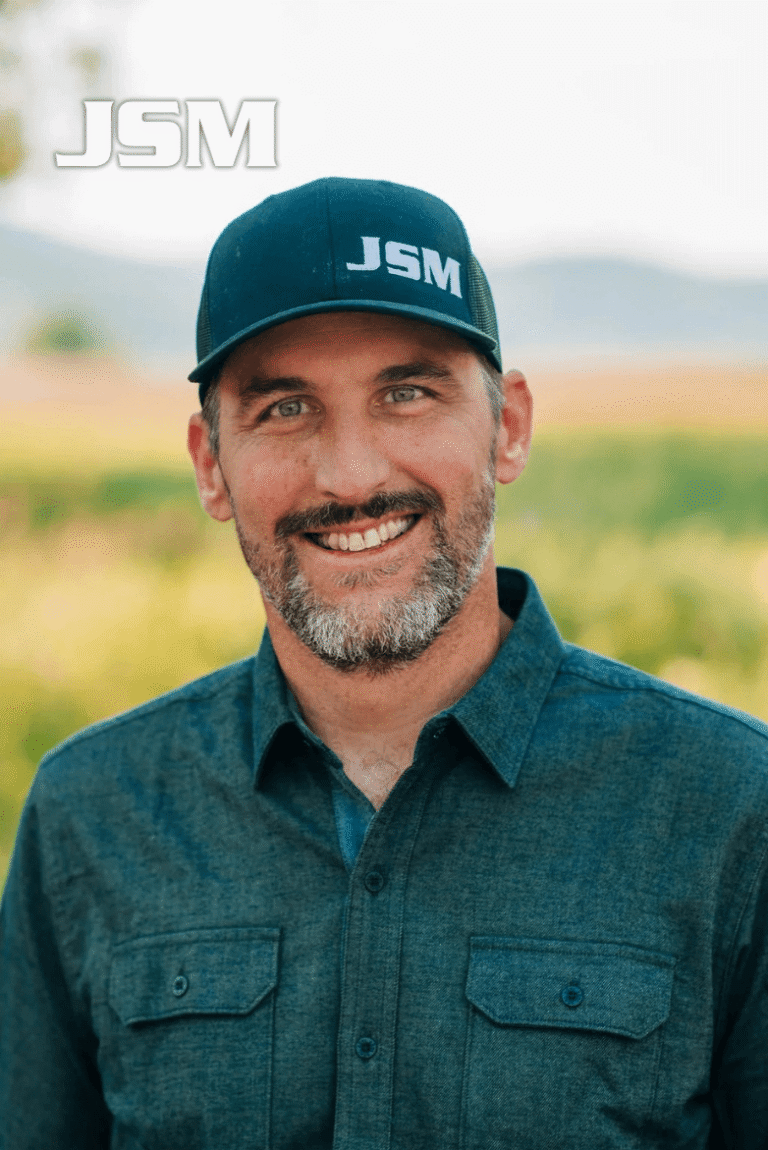 Portrait of Jeremy MacGray, President of JSM, smiling outdoors in a blue shirt and JSM hat.