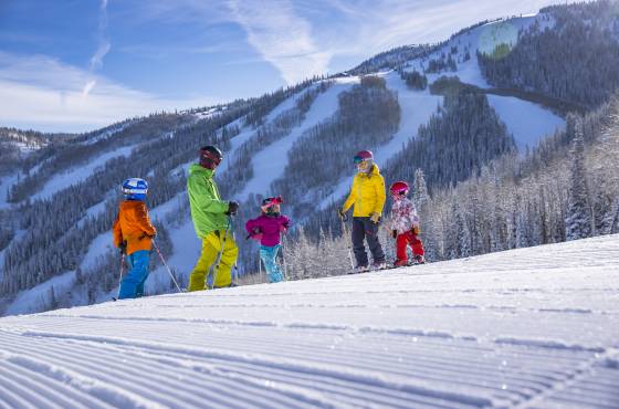 A family with children skiing on a freshly groomed slope at Steamboat Ski Resort under a blue sky, with snow-covered trees and mountains in the background.