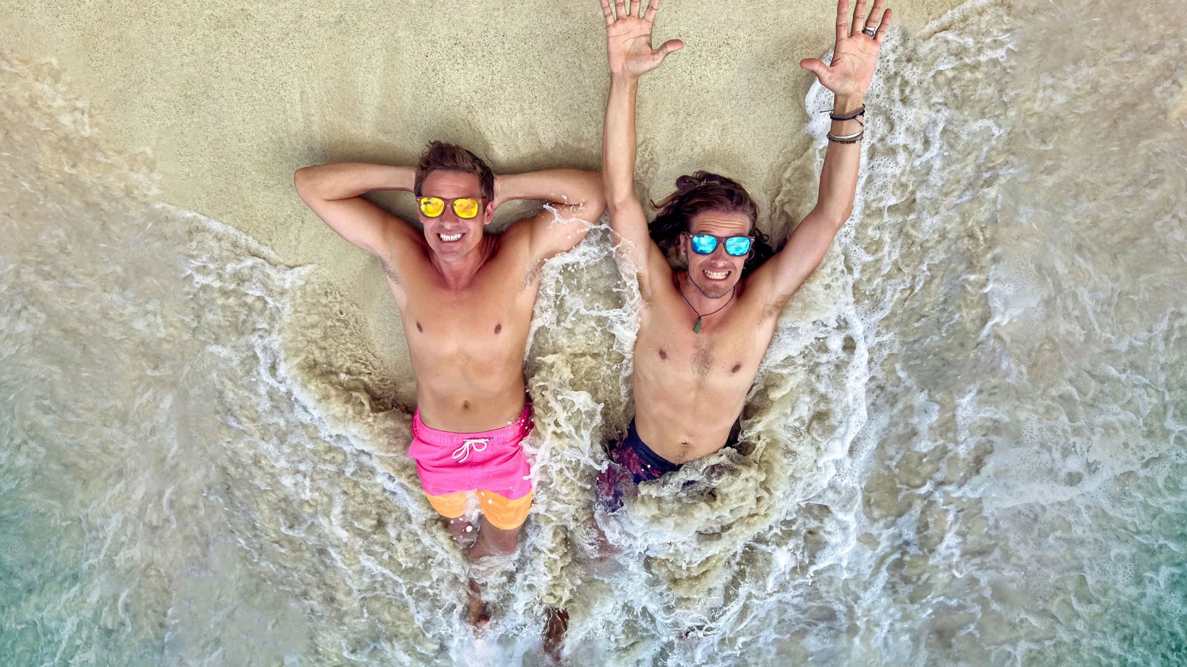 Two men smiling and lying in shallow surf with arms raised and sunglasses on, enjoying the beach and waves.